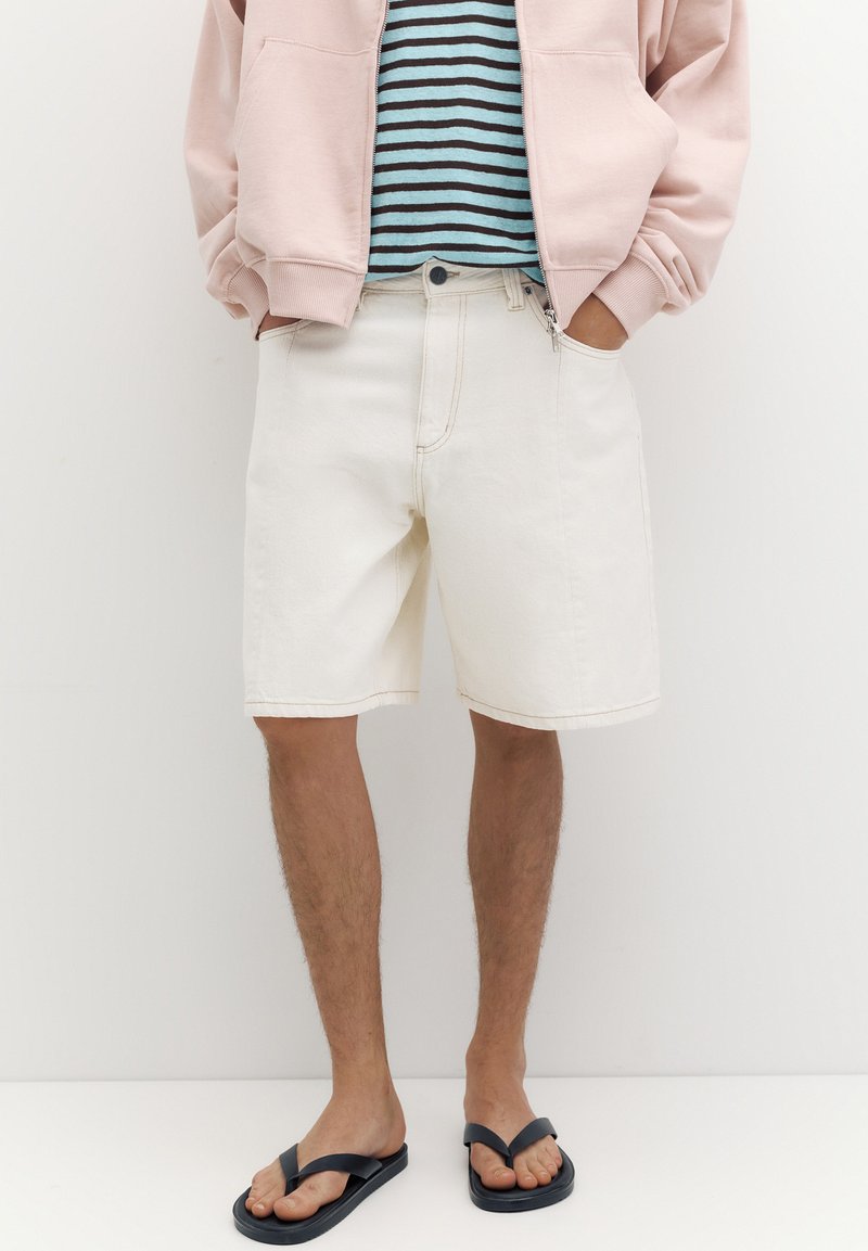Personne portant une veste rose, une chemise rayée bleue et noire, un short blanc jusqu’aux genoux et des tongs noires, les mains dans les poches, debout.