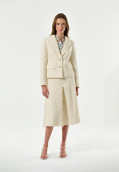 Costume texturé de couleur crème avec une veste ajustée à deux boutons et poches, associé à une jupe de longueur genou avec une fente.