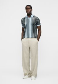 Mann trägt ein blau-schwarz vertikal gestreiftes Poloshirt, beige weite Hose und weiße Sneakers, steht mit den Händen in den Taschen.