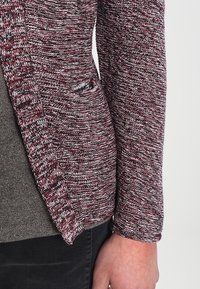 Pull en maille bordeaux et gris avec une surface texturée, doté d'une poche latérale unique et d'une coupe ajustée. La manche comprend un détail de poignet.