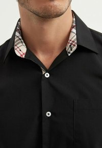 Chemise noire à boutons avec un col à motif tartan beige, rouge et noir, dotée de boutons blancs et d'une poche poitrine.