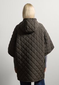 Cappotto trapuntato in olivo scuro con cappuccio, caratterizzato da un motivo a diamante. Vestibilità ampia con maniche corte, indossato sopra un maglione grigio e jeans blu.