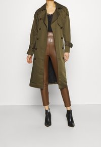 Trench coat verde oliva con bottoni doppi, maniche lunghe e vita con cintura, abbinato a pantaloni marroni fitted e stivali neri alla caviglia.