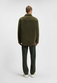 Olivgrüne Fleecejacke mit einem Kragen-Design, strukturiertem Stoff und geradem Schnitt. Kombiniert mit dunkelgrünen, geraden Hosen und weißen Schuhen.