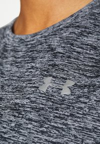 Detail osoby oblečené ve šedém sportovním tričku s texturou a reflexním logem Under Armour na hrudníku.