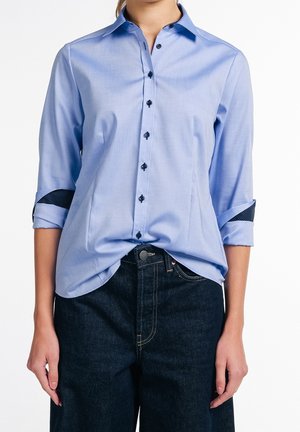 Frau trägt ein hellblaues Hemd mit Knöpfen und hochgekrempelten Ärmeln, die gemusterte Innenmanschetten zeigen, leicht in dunkelblaue Jeans gesteckt.