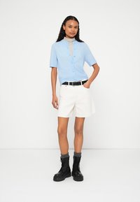 Gaiši zils īsroku kardigans ar pogām priekšpusē, nēsāts virs svītrainas augstas apkakles džempera. Kombinēts ar baltām šortiem un melniem kaujas zābakiem.