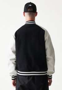 Μαύρο και άσπρο bomber μπουφάν με μαλακό μάλλινο πίσω μέρος, δερματίνη στα μανίκια, ριμπ γιακά και ριγέ μανσέτες. Λείο υλικό, κλασικός σχεδιασμός.