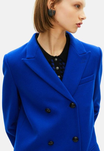 Kobaltblauer, doppelt geknöpfter Wollmantel mit Reverskragen, schwarzen Knöpfen und einer Brusttasche. Darunter ein strukturierter schwarzer Pullover.
