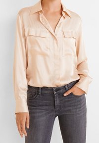 Blusa de seda crema con mangas largas, cierre de botones en la parte delantera, dos bolsillos en el pecho y un cuello clásico, combinada con jeans gris oscuro.