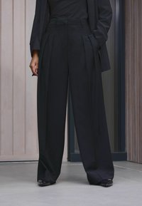 Pantaloni neri a gamba ampia con pieghe frontali, vita alta e tessuto liscio. Indossati con un top aderente e calzature nere.