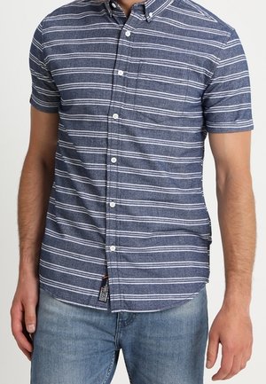 Man draagt een navyblauw overhemd met korte mouwen met witte horizontale strepen en lichtblauwe jeans.
