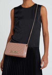 Průsvitná růžová kabelka přes rameno s dekorativním prošíváním a zlatými prvky, s logem a řetízkovým popruhem, nošena s černým bezrukávovým topem a skladanou sukní.