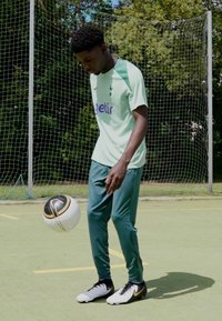 Zaļa sporta apģērba komplekts, kas sastāv no īsām piedurknēm augšpusē un sašaurinātiem biksēm, ar melnbalto futbolu, kas turēts tuvu kājām. Ārpus telpām.