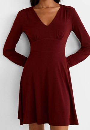 Kobieta ubrana w głęboko czerwoną sukienkę z długim rękawem, dekoltem w serek, dopasowaną w talii i rozkloszowaną spódnicą, stoi na jednolitym tle.