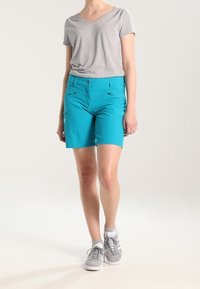 Tyrkysové šortky s hladkou texturou, vybavené kapsami na zip, spárované se světle šedým tričkem a šedými teniskami.