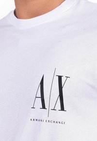 White cotton t-shirt featuring a black logo with "A/X" and "ARMANI EXCHANGE" text. Round neckline with a soft texture.