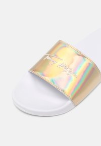 Sandale slide blanche avec une large bride dorée irisée arborant la signature Tommy Hilfiger en écriture blanche.