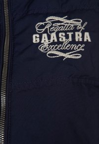 Granatowa kurtka na zamek z wyszywanym białym napisem "Regatta of GAASTRA Excellence", o gładkiej fakturze tkaniny.