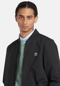 Giubbotto bomber nero con una texture liscia e colletto a coste, caratterizzato da un piccolo logo ricamato sul petto, indossato sopra un maglione verde e una camicia blu.