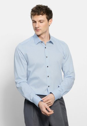 Man with short curly hair wearing light blue patterned button-up shirt and gray trousers, adjusting his cuff, looking down left.