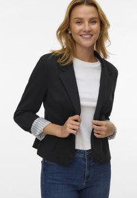 Schwarze Blazer mit figurbetontem Design, ausgestattet mit hochgerollten gestreiften Ärmeln, getragen über einem weißen T-Shirt und kombiniert mit blauen Jeans.