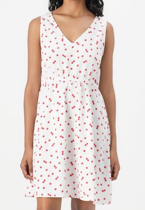 Ærmeløs hvid kjole med rødt kirsebærmønster, V-udskæring og samlet talje, båret af person med armene afslappet ned langs siden.