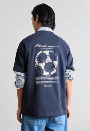 Muž nosi tmavomodré tričko s textom škandinávskej futbalovej komunity a grafikou vintage futbalovej lopty na chrbte.