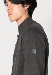 Grijze zip-up truien met ribbelkraag en -manchetten, voorzien van een gestructureerd breipatroon en een klein zwart leren logo-opdruk op de schouder.