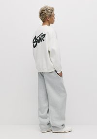Sweatshirt blanc surdimensionné avec du texte noir dans le dos, associé à un pantalon de survêtement gris ample et des baskets blanches. Le modèle se tient de profil.