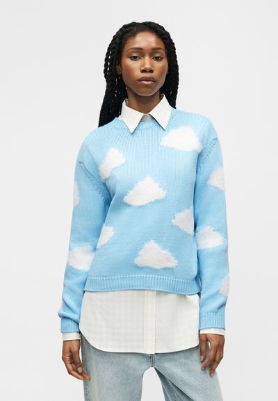 Femme aux longs cheveux tressés portant un pull bleu clair avec des nuages blancs, une chemise blanche à col et un jean bleu clair, sur fond gris.