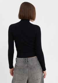 Pull noir en maille côtelée avec col montant et manches longues, associé à un jean gris délavé taille haute avec poches arrière.
