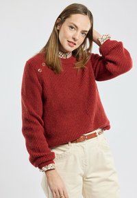 Pull en maille côtelée rouge avec encolure ronde, comportant deux boutons décoratifs sur l'épaule, porté sur une chemise à carreaux et un pantalon beige.