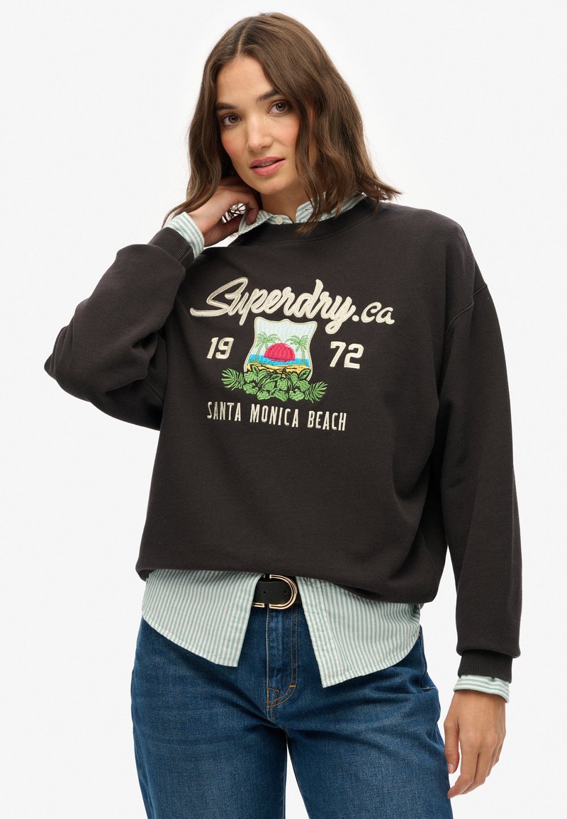 Czarna, krótka bluza z haftem "Superdry.ca" oraz "Santa Monica Beach", założona na jasnoniebieską koszulę w paski, w połączeniu z niebieskimi dżinsami.