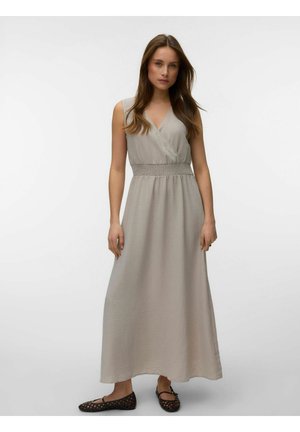 Kobieta stoi, ubrana w bezrękawnikową, jasnobeżową maxi sukienkę z dekoltem w serek i elastyczną talią, w parze z czarnymi plecionymi balerinami.