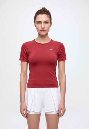 Une femme se tient face à l’objectif devant un fond uni, vêtue d’un maillot de sport rouge à manches courtes et d’un short de sport blanc.