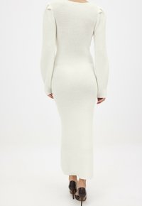 Robe longue et ajustée avec une texture côtelée, de couleur blanche, et des manches larges, associée à des chaussures à talons foncés.