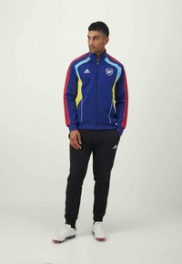 Modrá bunda na zips s červenými pruhmi a viacfarebnými akcentmi, spárovaná s čiernymi nohavicami a bielou obuvou. Viditeľné logo Adidas.