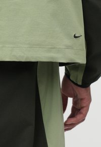 Světle zelený svetr s tmavě zeleným akcentem, s malým černým logem Nike a rovné lemování. Hladký materiál a vrstvený design.