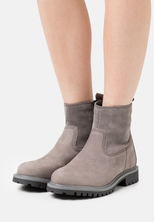 Ein Paar graue Wildleder-Ankle-Boots mit dicken schwarzen Sohlen, getragen an hellhäutigen Beinen vor einem weißen Hintergrund.