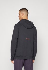 Sweatshirt à capuche gris foncé en tissu doux. Présente un logo Reebok orange au dos et des poignets côtelés. Design et coupe simples.