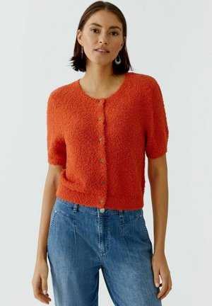 Vrouw draagt een getextureerd, oranje, kort mouwtjes, doorgestikt vest met knopen en een high-waisted blauwe jeans tegen een effen achtergrond.