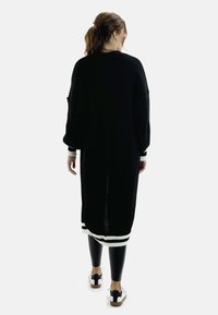 Suéter largo negro de punto con mangas oversized, puños y dobladillo de canalé blanco, combinado con leggings negros y zapatillas blancas.
