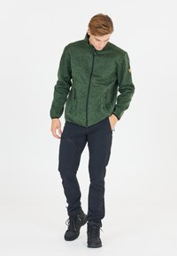 Grön texturerad fleecejacka med hög krage, framzip och två sidfickor, i kombination med svarta byxor och svarta skor.