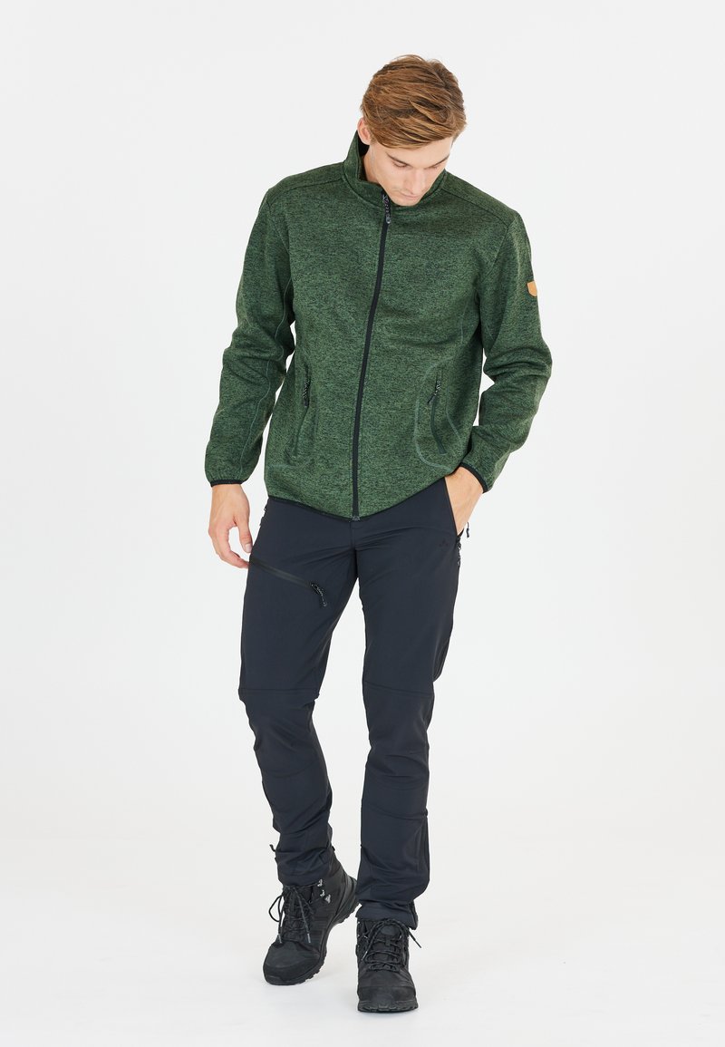 Grön texturerad fleecejacka med hög krage, framzip och två sidfickor, i kombination med svarta byxor och svarta skor.