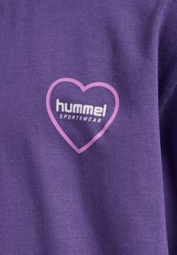 Lilla bomulds-T-shirt med en hjerteformet pink kant og teksten "hummel" samt "SPORTSWEAR" i hvid. Teksturdetalje i stoffet er synlig.