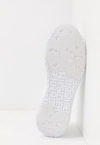 Valkoinen lenkkarin pohja, jossa on kohotettu logo-kuvio. Sileä pinta ja pyöristetyt reunat, jotka näyttävät kuvion gripin vuoksi. Pystysuunnassa.