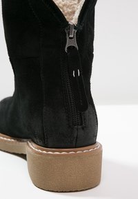 Bottine en daim noir avec une fermeture éclair, intérieur doublé, semelle texturée couleur tan et détails cousus autour de la semelle extérieure.