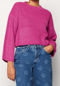 Magenta stickad tröja med rund hals och vida ärmar kombinerad med blå denimjeans med ett mönster av peace-tecken.