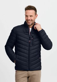 Svart polstret jakke med høy krage, med horisontalt quiltet design, glidelåslomme på brystet og kontrastfarger på glidelåsene.